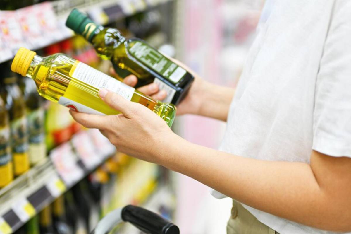 A shopper compares two bottles of oil in a grocery store, holding a green and a yellow bottle while examining the labels.