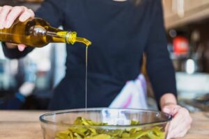 A person in a dark shirt pours olive oil from a glass bottle into a bowl of green vegetables on a kitchen countertop.